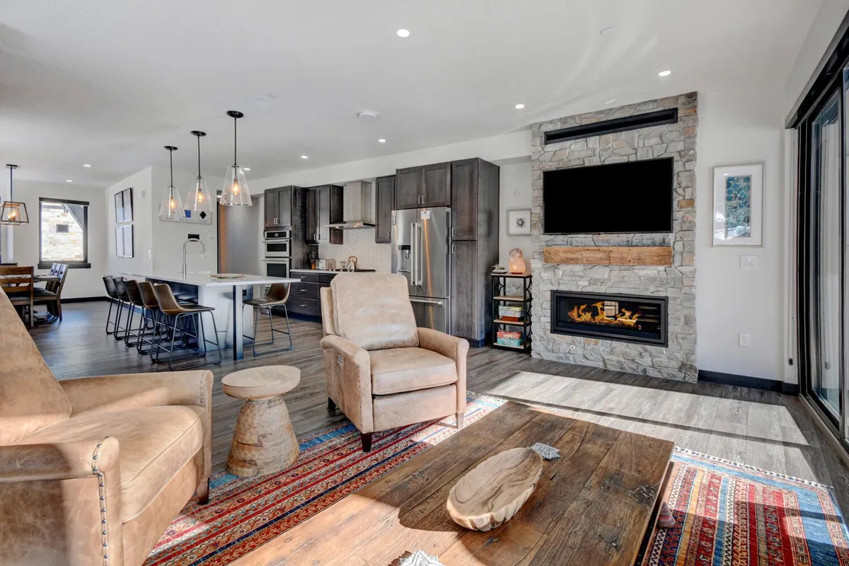 An inviting open-concept living space featuring a cozy fireplace, a modern wooden coffee table, a large dining area, and a kitchen island in the background. The arrangement of comfortable seating and warm lighting suggests a perfect environment for social gatherings, family movie nights, and shared meals.