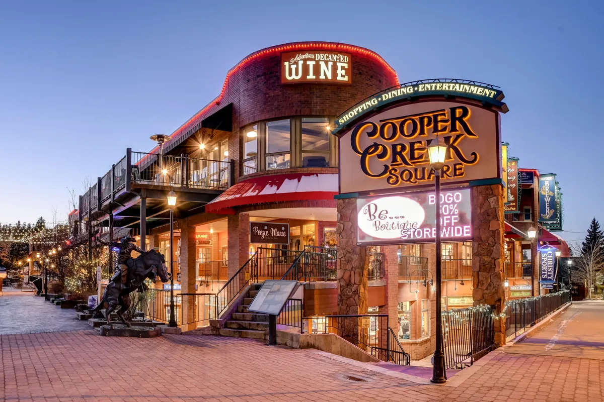 This exterior image captures the lively Cooper Creek Square in downtown Winter Park, featuring a prominent sign, a wine shop, a bustling outdoor dining area, and a distinctive horse sculpture. The scene is vibrant and welcoming, with visible activity and hints of local entertainment and shopping options.