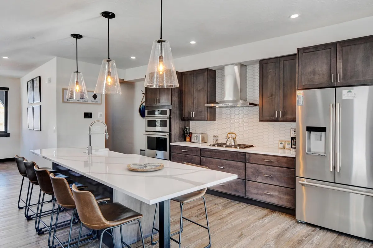 A spacious, contemporary kitchen featuring a large island countertop with pendant lighting above, sleek stainless steel appliances, and generous workspace ideal for meal preparation and entertaining. The design exudes warmth with its mix of modern finishes and inviting atmosphere, suggesting ample room for both cooking and gathering.