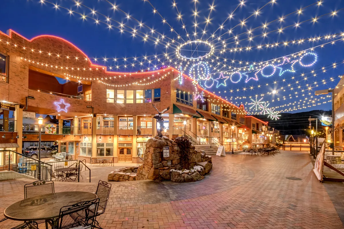 An inviting view of a downtown plaza at night, decorated with festive lights, a central fountain, outdoor seating, and illuminated storefronts. People can be seen engaging in evening dining and social activities, creating a sense of community and celebration.