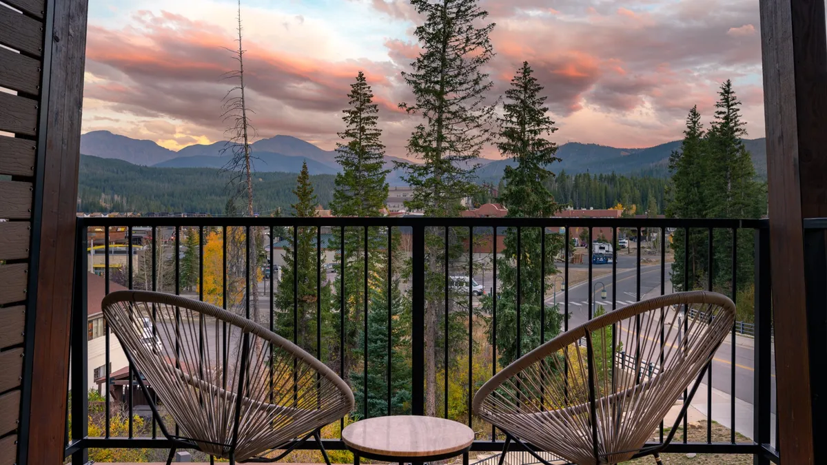 An inviting balcony overlooks a breathtaking mountain landscape, framed by lush trees and drifting clouds. The outdoor seating on the balcony offers a peaceful place to relax and take in the scenery, suggesting a seamless connection between comfort and nature.