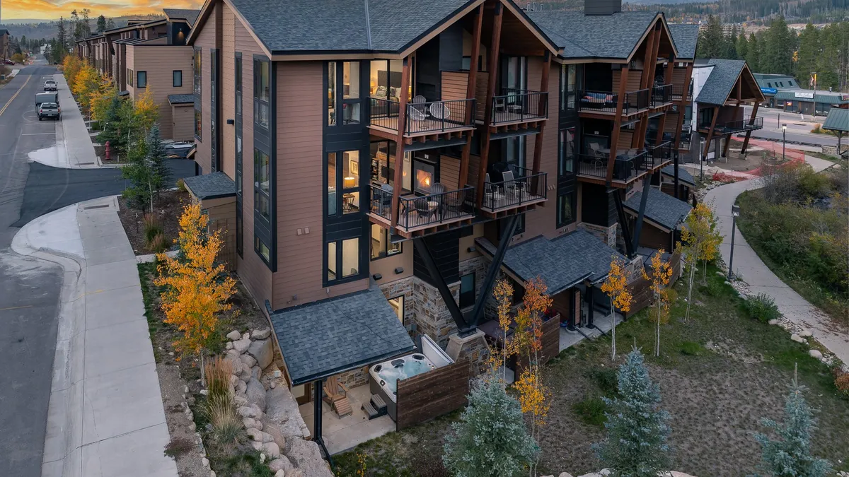 The image showcases the exterior of a luxurious single-family house nestled among tall trees in Winter Park, Colorado. Spacious balconies overlook a stunning riverfront landscape, while a private hot tub is perfectly situated for relaxation. An inviting asphalt pathway winds through the property, hinting at memorable strolls and easy outdoor access. The setting is awash in the warm glow of sunset, creating a tranquil and welcoming atmosphere.