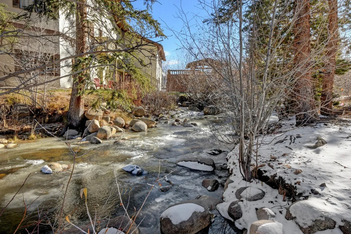 A tranquil view of a meandering stream flanked by snow-covered rocks and trees, with the building’s facade visible nearby. The scene exudes peace and natural beauty, inviting guests to enjoy serene walks along Vasquez Creek and experience the outdoors year-round.