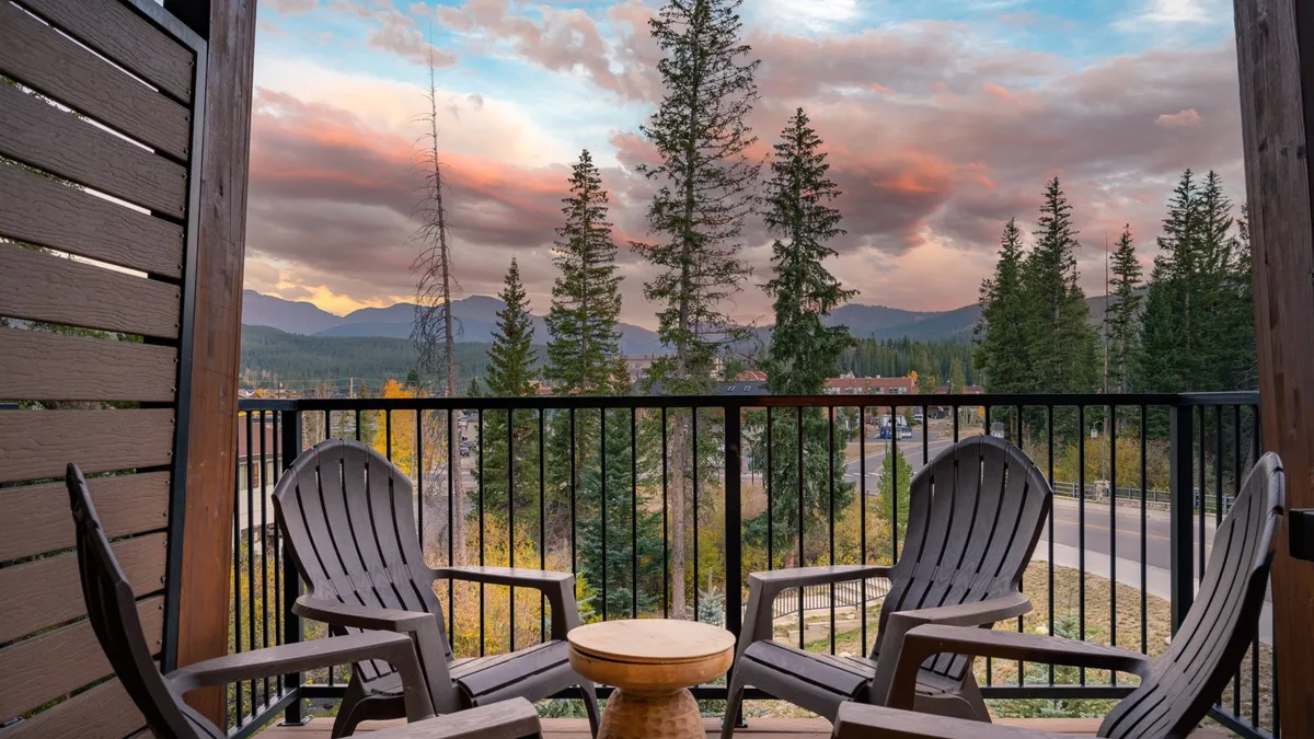 Wooden chairs and a small table are placed on a deck overlooking a dramatic mountain view, with a glowing sunset sky and evergreens in the distance. The scene is peaceful and designed for morning coffee or evening relaxation.