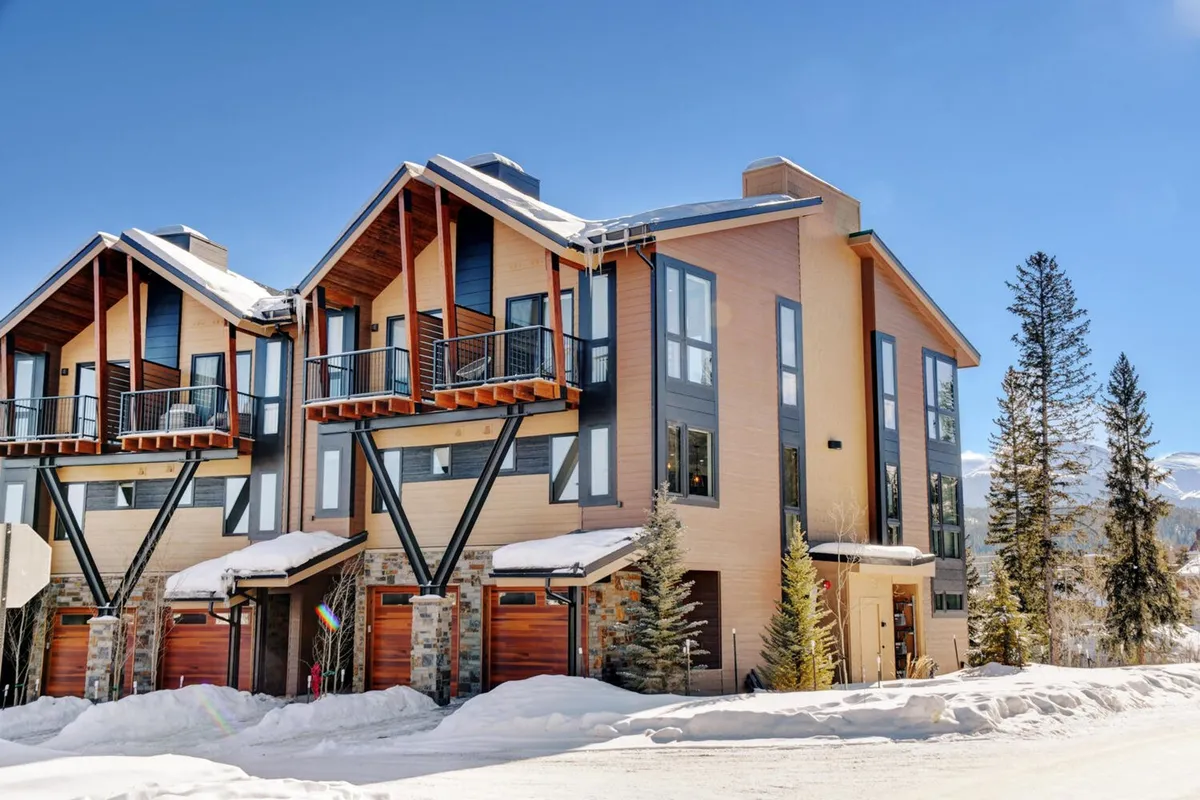 An inviting exterior shot showing modern mountain architecture with stone accents, balconies, and a snow-covered landscape. Guests can be seen enjoying activities like skiing and snowboarding, with the resort ambiance and mountain views setting the scene.