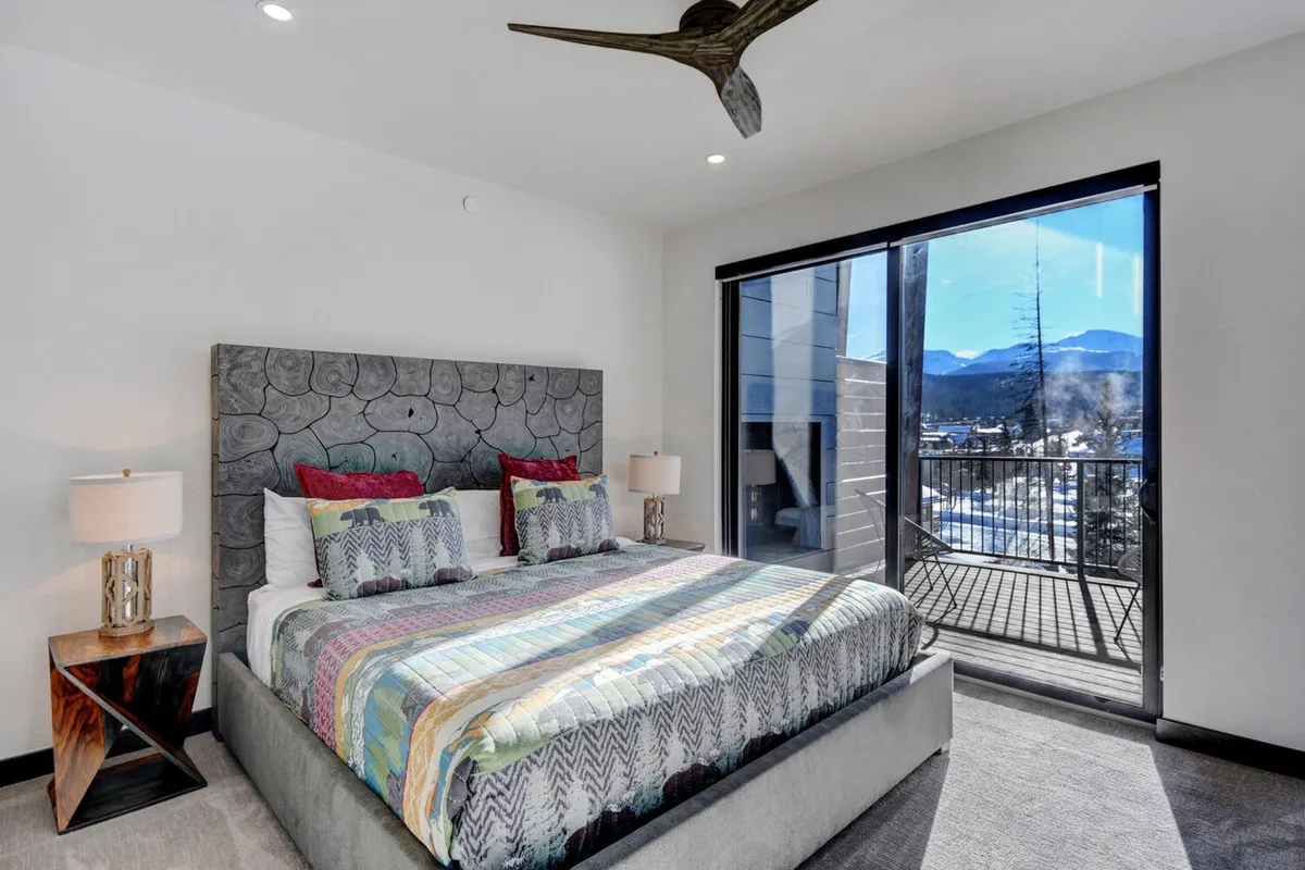 A serene bedroom featuring a bed with colorful bedding, a textured headboard, and expansive windows offering a stunning mountain view. The space exudes calm and comfort, with natural light streaming in and a cozy atmosphere ideal for rest.