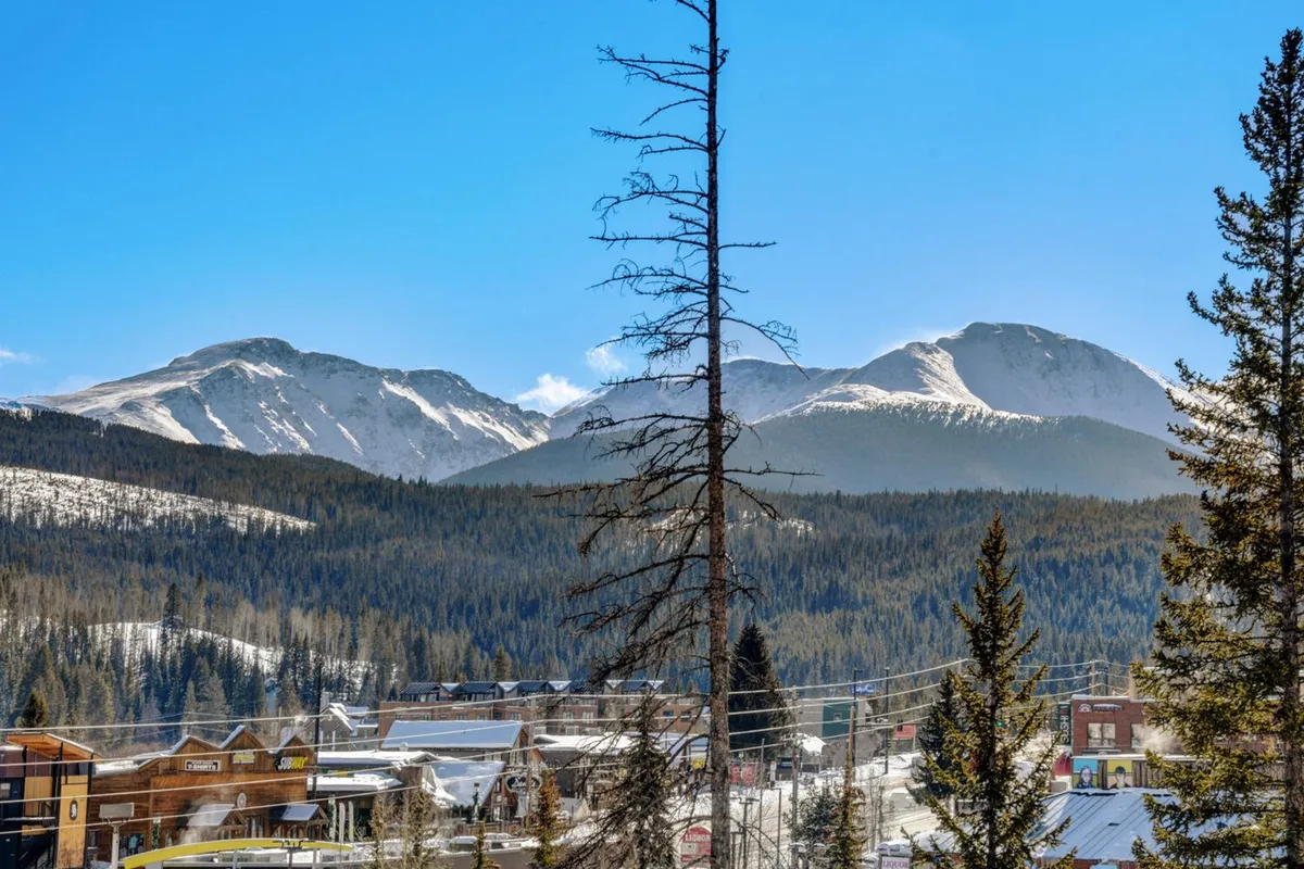 A panoramic view captures majestic mountains, pine forests, and the charming town below. The scene is tranquil and expansive, offering a sense of escape and highlighting the region’s outdoor appeal and beauty.