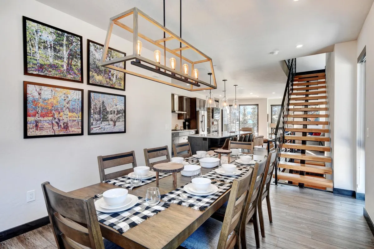 A large, elegant dining table set for a gathering, framed by artistic wall décor and a striking lighting fixture. The space is open and inviting, with a staircase in the background suggesting a connection to other living areas. The ambiance is warm, ideal for family dinners or elaborate celebrations.